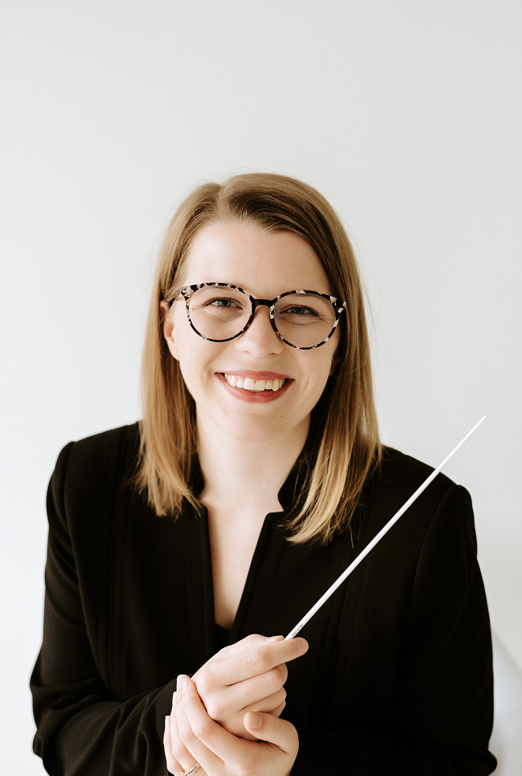 Portrait of Danielle Bridges, a young woman with light brown hair past her shoulders, wearing a black blazer and holding a conducting baton. She is wearing glasses and is smiling
